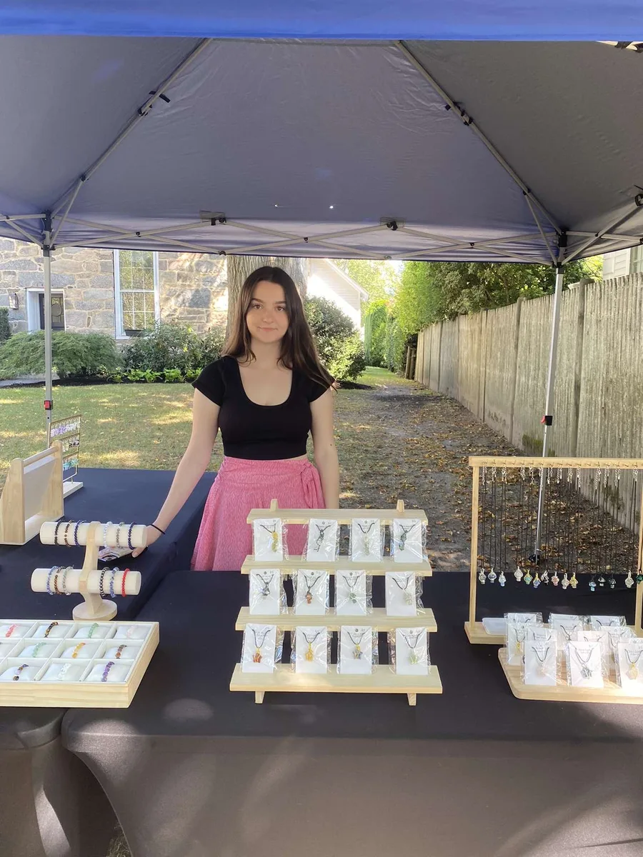 Alyssa standing under a blue canopy at her crystal jewelry booth, with necklaces and bracelets on wooden stands
