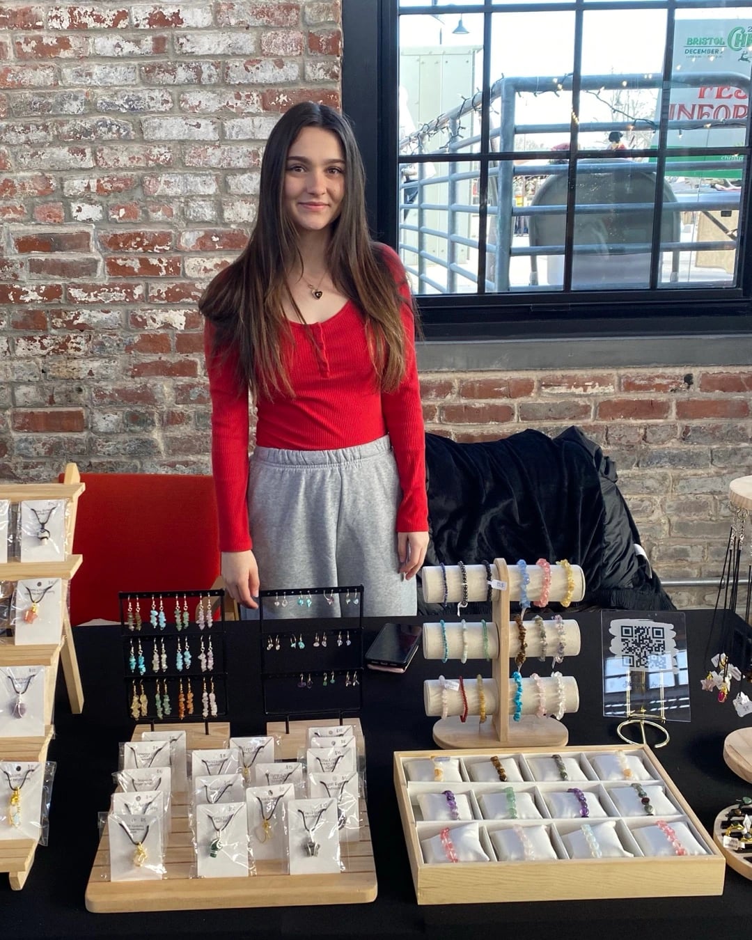 Alyssa behind her jewelry table at a market, with earrings, bracelets and necklaces on display